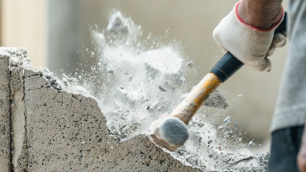 Ein Arbeiter schlägt mit einem Hammer auf eine Betonwand, während Staub und kleine Steine in die Luft fliegen.