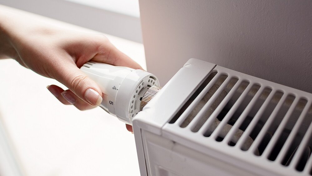 Eine Hand dreht den Thermostat einer Heizung, um die Temperatur in einem hellen Raum zu regulieren.