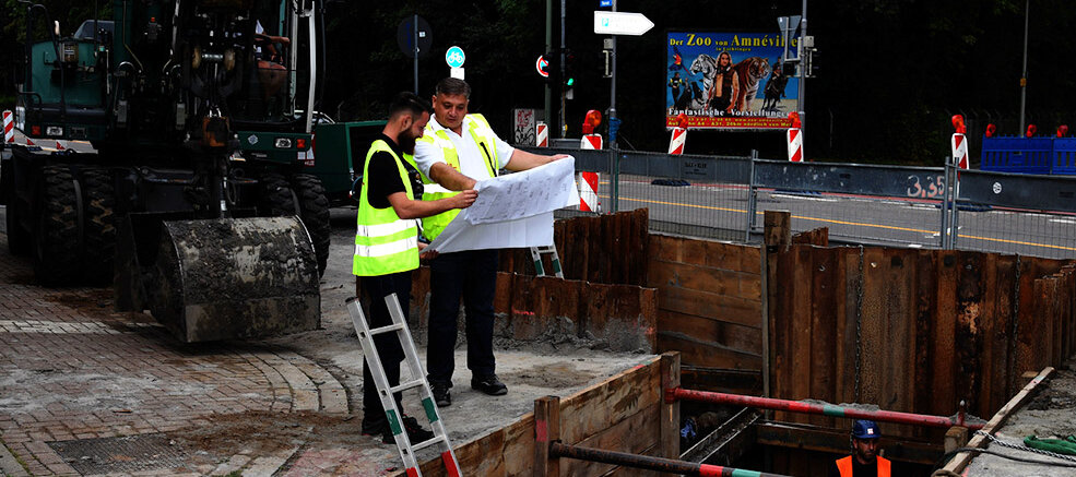 Zwei Bauarbeiter in Warnwesten studieren Pläne über einer Baustelle mit einem großen Bagger im Hintergrund.