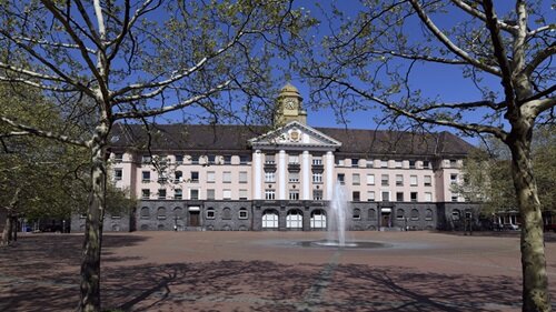 Ein historisches Gebäude mit einer beeindruckenden Fassade und einem zentralen Brunnen, umgeben von Bäumen im Frühling.