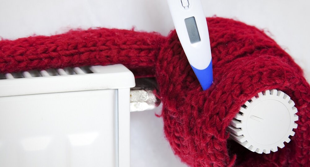 Ein roter Wollschal wickelt sich um einen Heizkörper, während ein Thermometer darauf liegt und die Temperatur anzeigt.