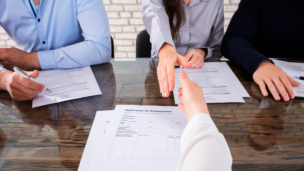 Eine Person reicht ihre Bewerbungsunterlagen über den Tisch, während andere Teilnehmer aufmerksam zuschauen.