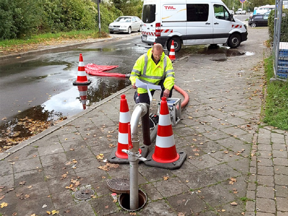 Ein Techniker arbeitet an einer Pumpe, um Wasser aus einem Gully zu entfernen, während Autos im Hintergrund parken.